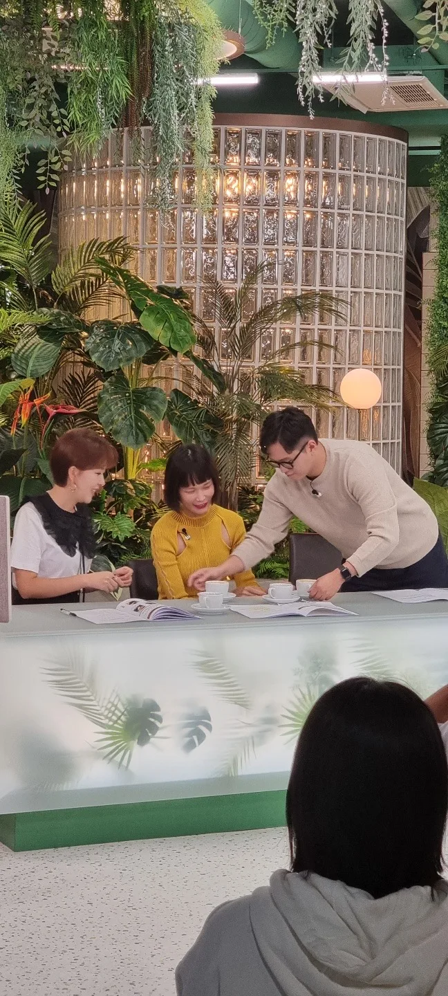 Three people are sitting at a table with white cups and papers, surrounded by lush green plants and foliage, in a cozy indoor setting.