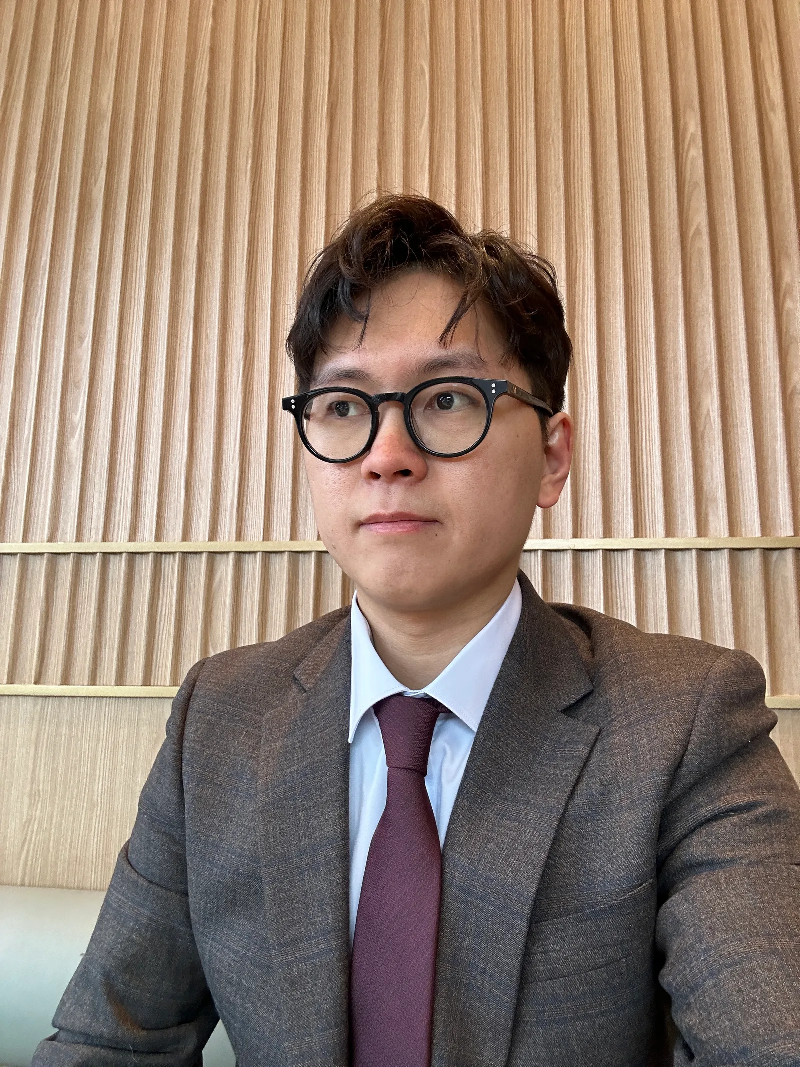 A man with dark hair and glasses wearing a brown suit, white shirt, and maroon tie, sitting in front of a wood-paneled wall.