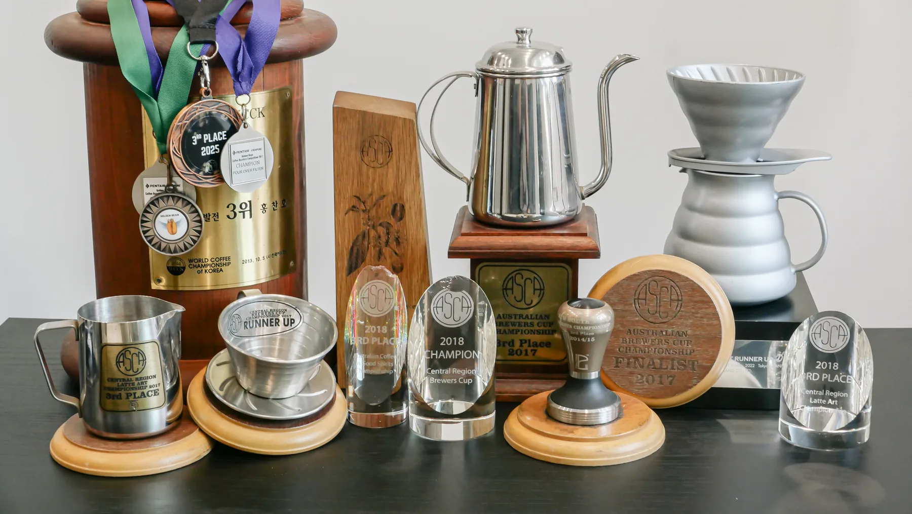 Collection of awards, trophies, and medals for coffee brewing competitions, along with a teapot, a dripper, and a milk pitcher displayed on a table.